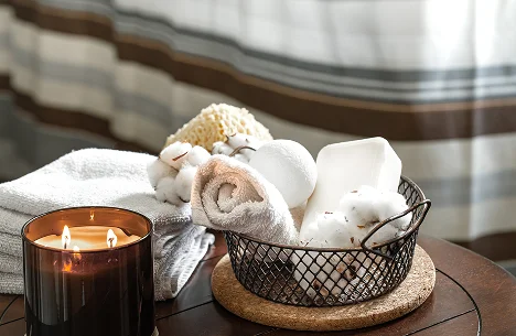 Een gezellige spa-scène met een brandende kaars, opgerolde handdoeken en badbenodigdheden in een mand op een houten tafel. Zacht licht zorgt voor een ontspannende sfeer.