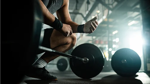Een man in sportkleding hurkt in een sportschool en houdt een gewichthefschijf vast bij een halterstang. De scène straalt focus en intensiteit uit, met een schemerige, sfeervolle achtergrond.